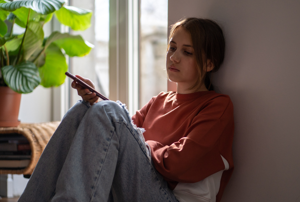 A teen girl appears alone and down while holding her phone, sitting by a window